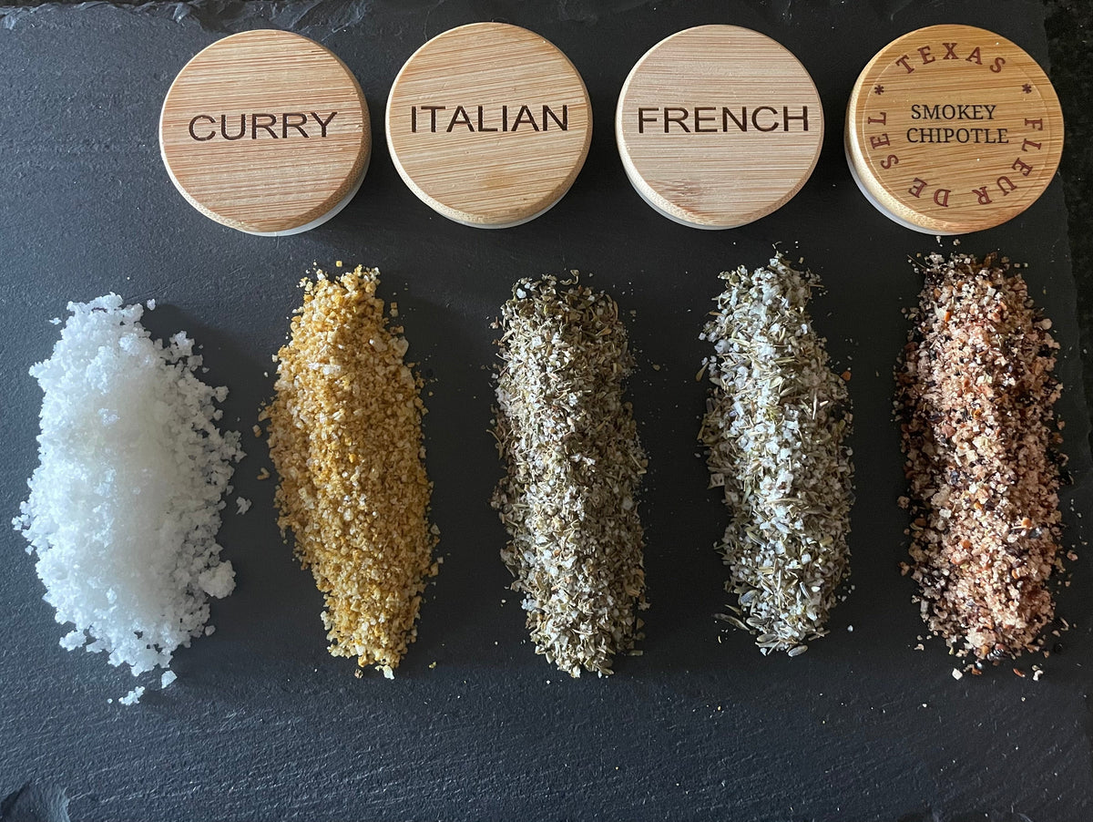 Four varieties of seasoning blends and Fleur de Sel salt on a dark slate background.