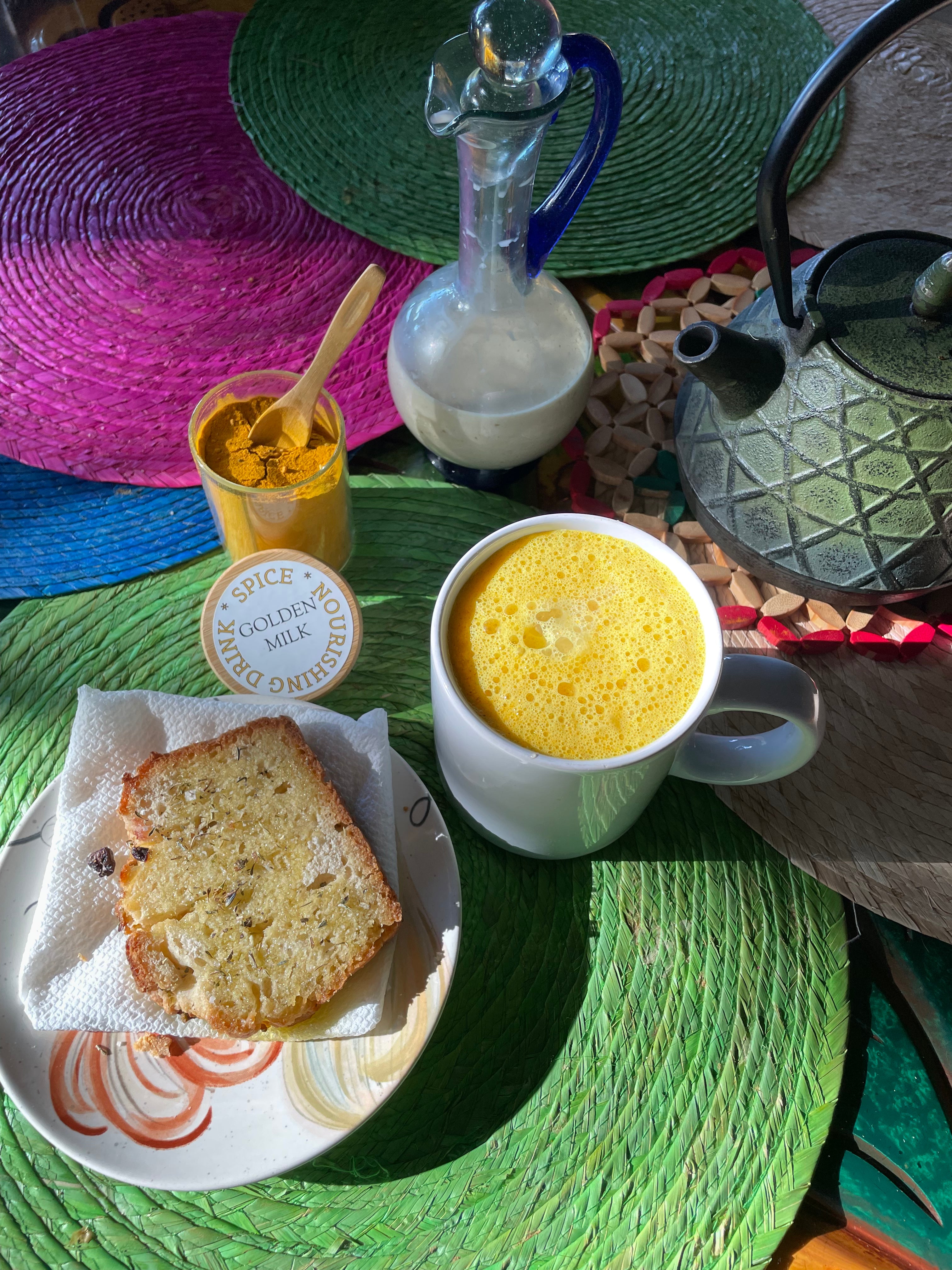 Golden turmeric latte, spice jar, glass jug of milk, and homemade bread on colorful placemats