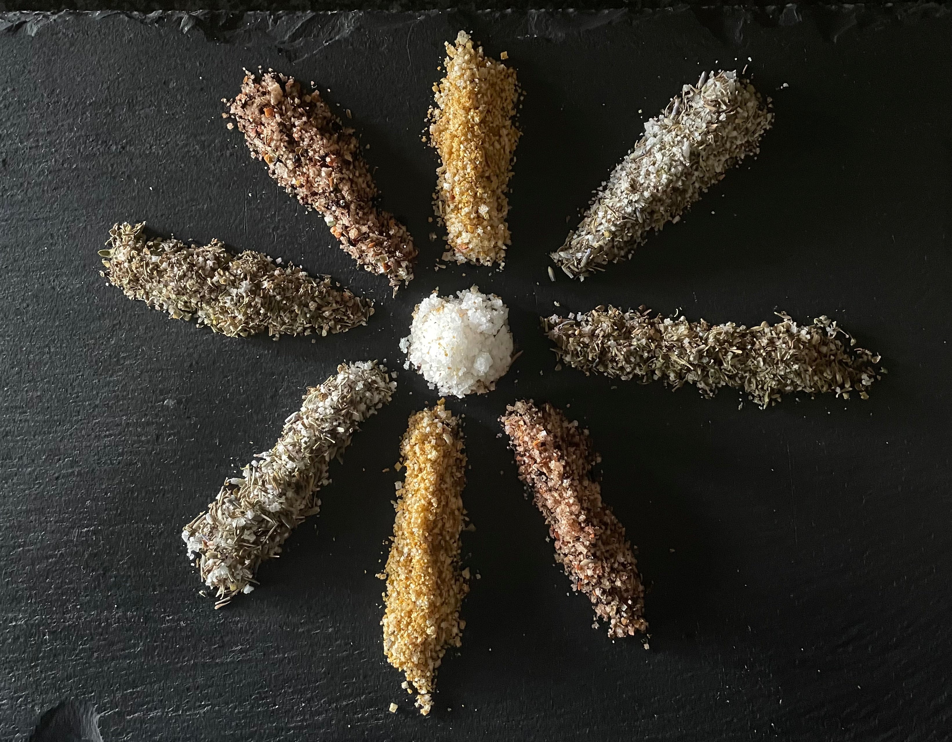 Assorted gourmet salts and dried herbs arranged in a sunburst pattern on black slate