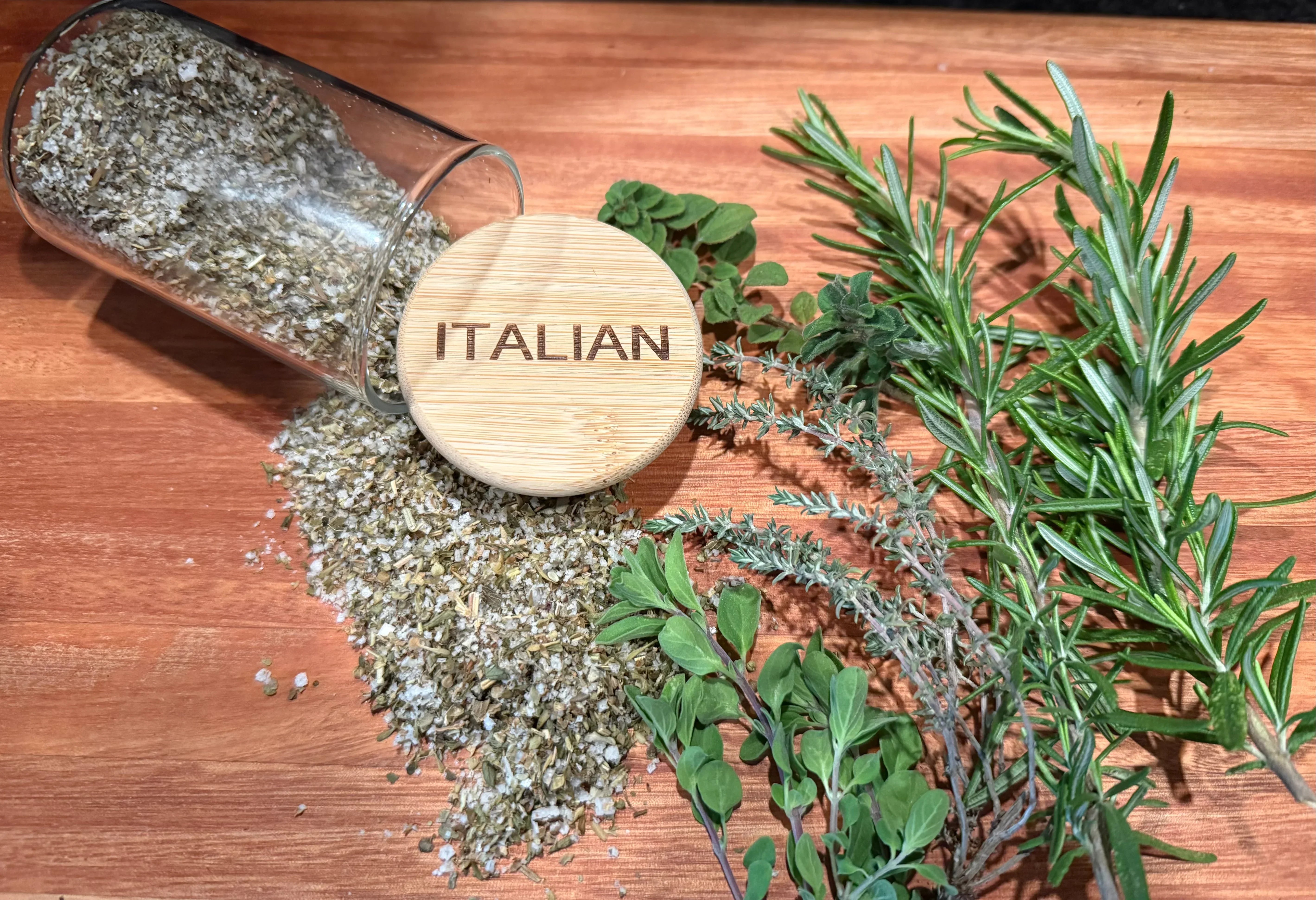 Italian herb salt blend spilling from glass jar with fresh rosemary, thyme, and oregano on wood