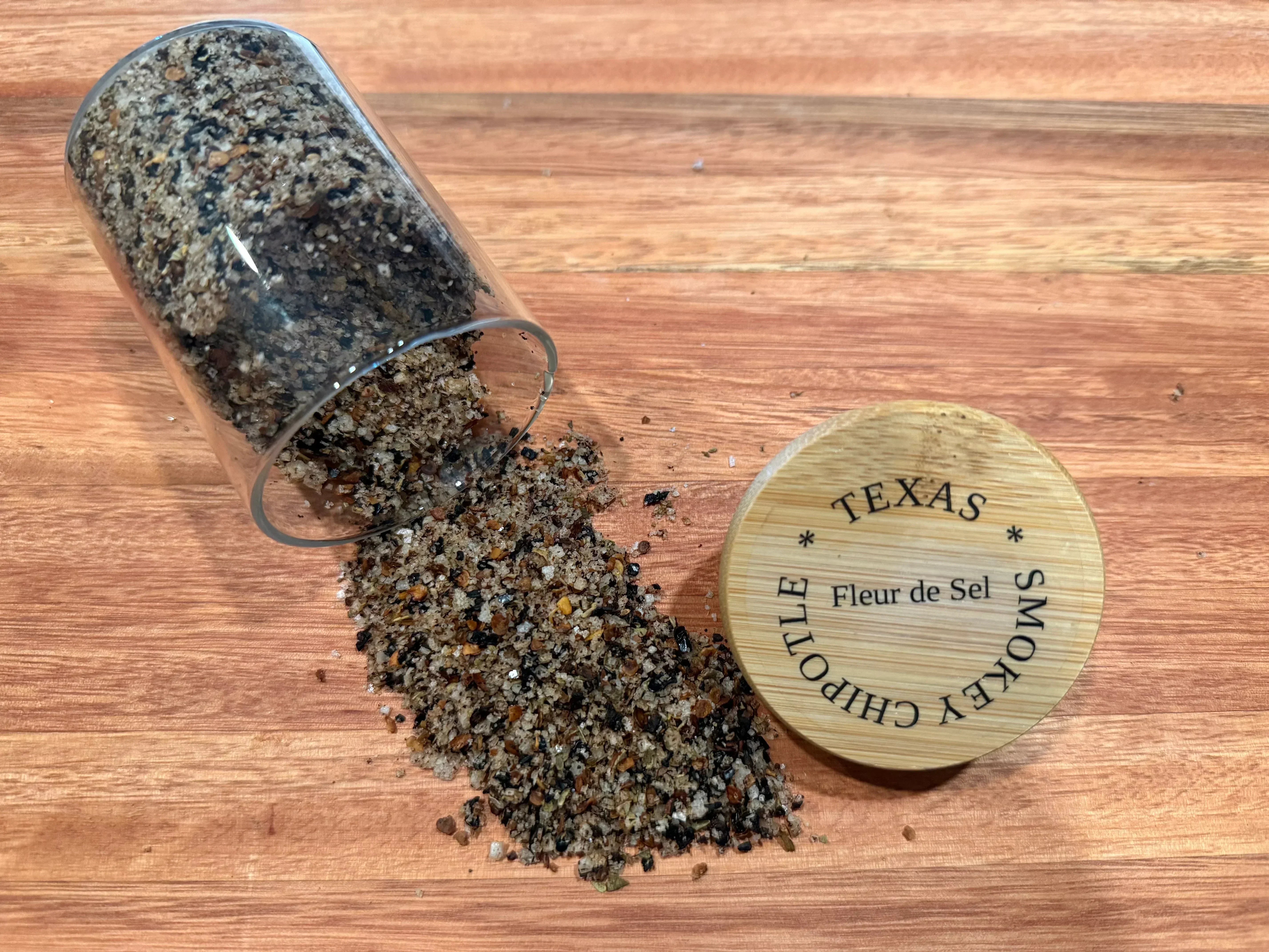 Glass jar spilling Texas Smokey Chipotle Fleur de Sel salt blend on wooden surface