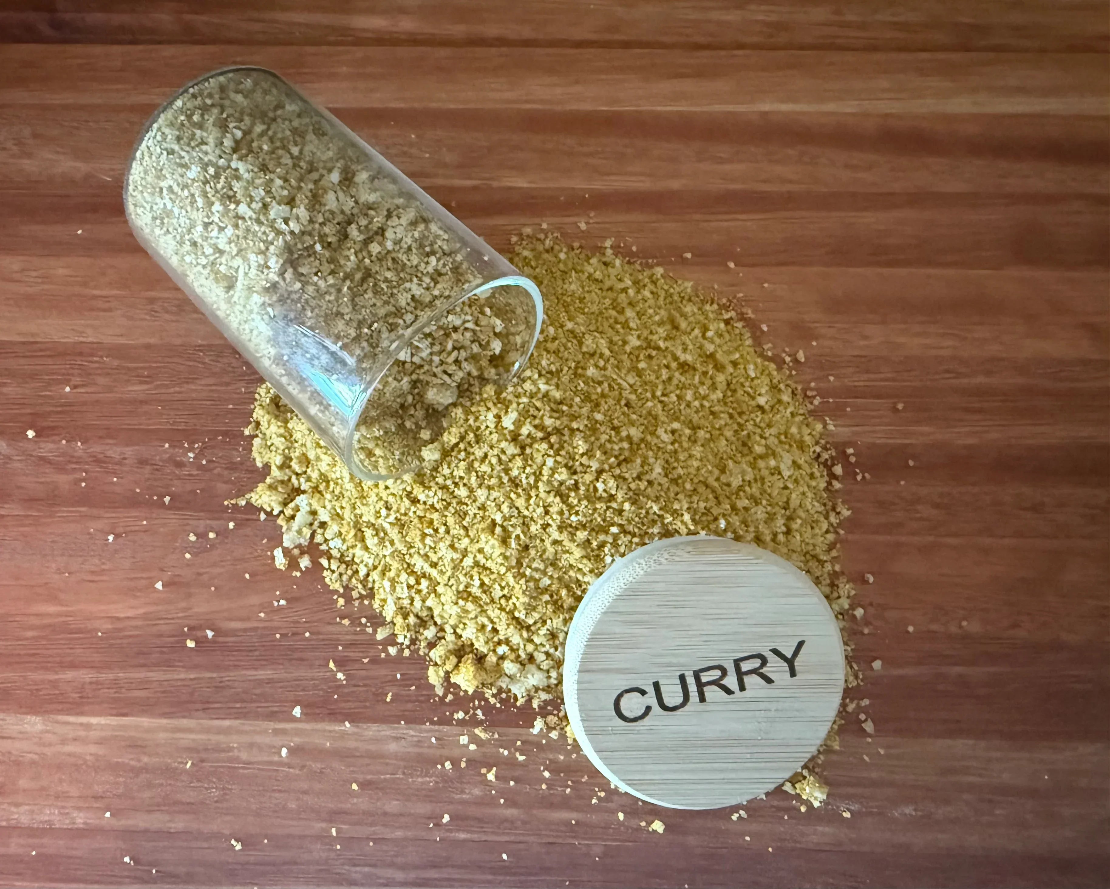 Spilled glass jar of curry fleur de sel salt on wooden surface with wooden lid labeled CURRY.
